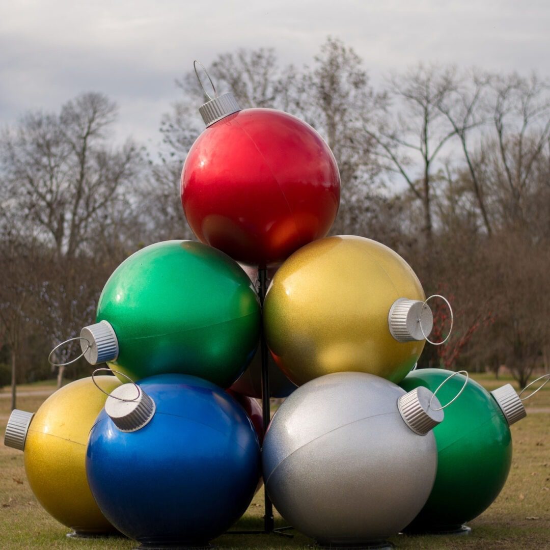 An image of ten (10) large multi-colored Christmas ornaments stacked on top of each other in a pyramid fashion. The ornaments are outside. There are six (6) ornaments on the bottom, four (4) ornaments in the middle, and one (1) ornament on the top.
