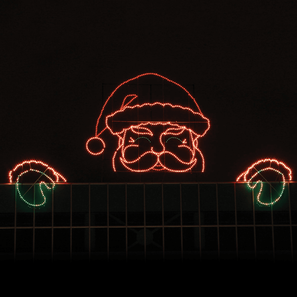 A gigantic illuminated Christmas silhouette of Santa peaking from a building roof with is hands on the edge of the roof.
