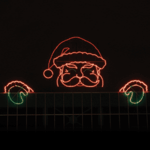 A gigantic illuminated Christmas silhouette of Santa peaking from a building roof with is hands on the edge of the roof.