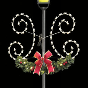 symmetrical, illuminated white scroll patterns on either side of the pole, decorated with small, warm white lights. Below, there is a green garland adorned with red ornaments and a large red ribbon with gold trim at the center. The background is dark, highlighting the warm glow of the lights.