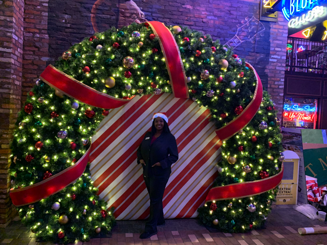 An image of a woman standing in front of a giant 9 foot, half wreath with a red and white stripped backdrop. The wreath is decorated with warm white C9 LED lights, ornaments, and a giant ribbon wrapping around the wreath.