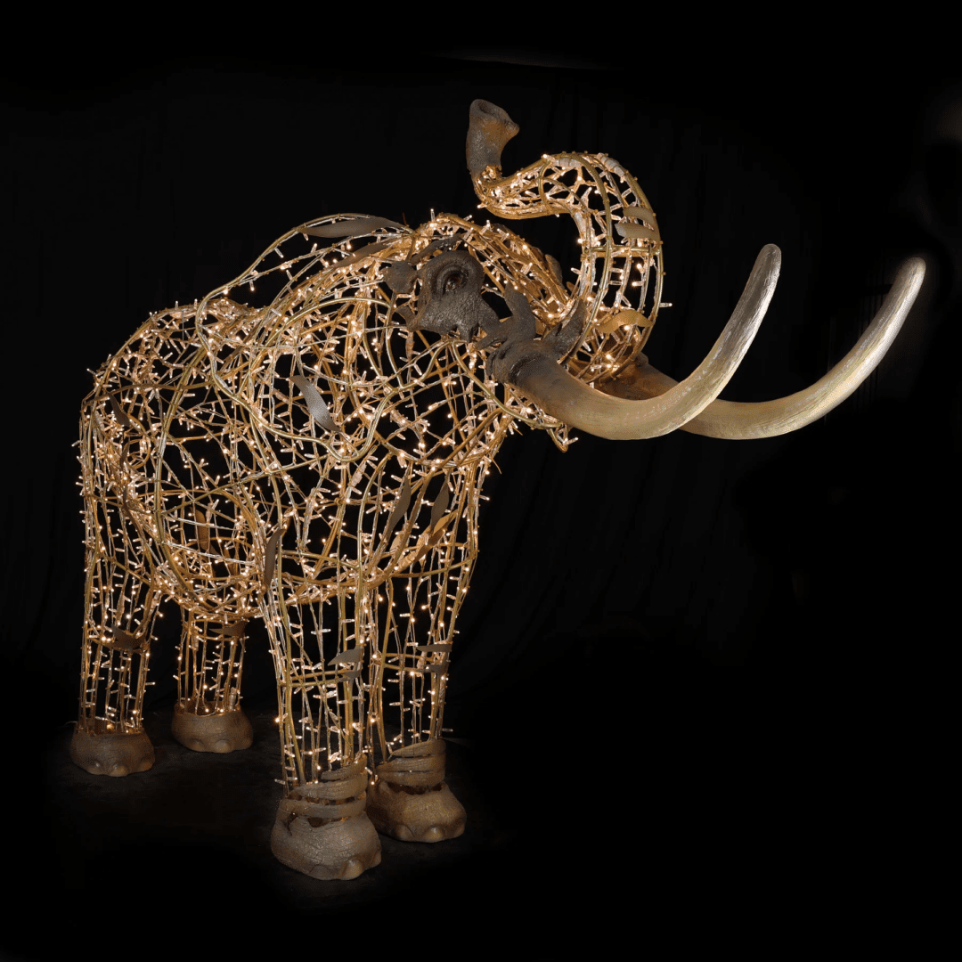 An image of a large elephant with long tusks. The elephant is made of a wire frame, warm white mini LED Christmas lights, and fiberglass accents.