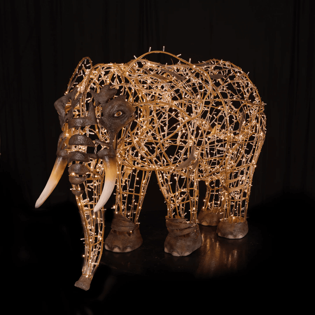 An image of a large elephant with long tusks. The elephant is made of a wire frame, warm white mini LED Christmas lights, and fiberglass accents.
