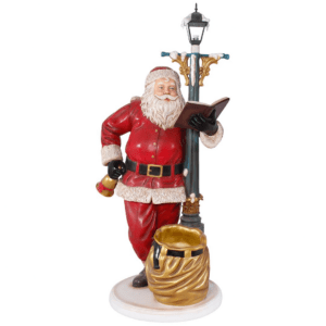 A fiberglass statue of Santa Claus leading against a lit lamp post. Santa is holding a bell and an open book.