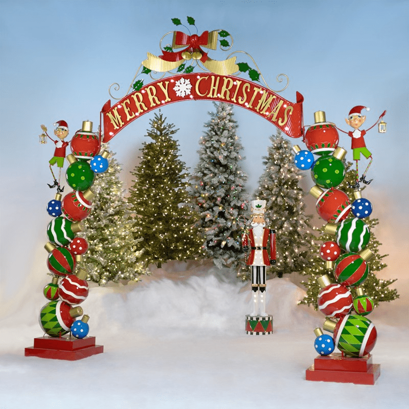 An image of a larch Christmas themed archway. The pillars are made up of stacked ornaments with an elf on each side. The top of the archway says "Merry Christmas" and has bells and holly leaves above it.