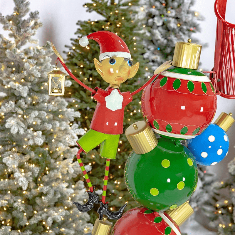 An image of a larch Christmas themed archway. The pillars are made up of stacked ornaments with an elf on each side. The top of the archway says "Merry Christmas" and has bells and holly leaves above it.