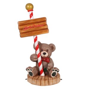 A brown teddy bear sitting down, holding a wooden sign on a red and white pole. The sign reads "Merry Christmas."